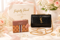 Black handbag with gold chain and brown wallet on a white surface with flowers and gift box in the background.